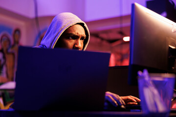 Focused african american scammer cracking password to gain access to system. Concentrated hacker in hood hacking database and breaching data in dark hideout place with neon light