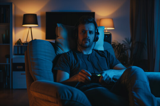 AI Generated Image. Serious man relaxing on a chair and playing video game while wearing headphones at night
