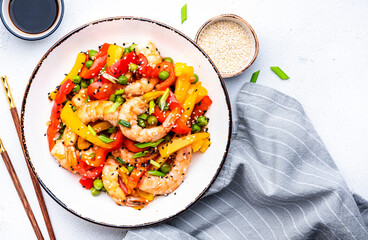 Spicy stir fry shrimps with colorful paprika, green peas, onion and sesame seeds with ginger, garlic and soy sauce in white bowl on kitchen table background, top view