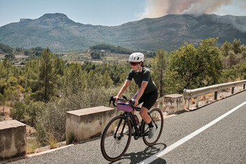 Woman cyclist riding a gravel bike with epic view of the Spanish mountains. Fit athlete wearing sportswear and helmet. Sports motivation image. Good road for cycling. Alicante region, Spain.