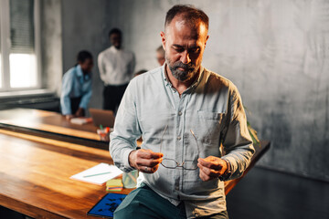 Mature businessman sits at boardroom and with eyeglasses in his hands.