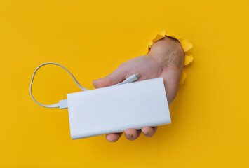 A right man's hand with a white metal power bank protrudes from a torn hole in yellow paper. Concept of smartphone charging and mobile energy independence.