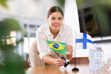 Positive young woman putting little flag of Brazil on the table with flag of Finland in conference...