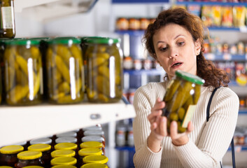 In Russian goods store, woman stand near showcase with canned vegetables, choose jar of pickled...
