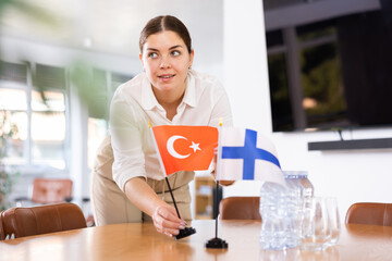Young female assistant setting up flags of finland and turkey on table for international...
