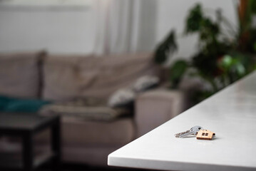 Keys with house shaped keychain on table on living room interior background. Concept of new homeownership, renting or leasing a property, and the process of purchasing a home, mortgage arrangements. - obrazy, fototapety, plakaty