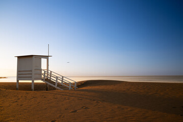 A small white building with a ladder leading up to it sits on a sandy beach. The sky is clear and blue, and there is a bird flying in the distance. The scene is peaceful and serene