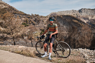 Portrait of a male cyclist in a helmet and cycling kit, standing near his bicycle on epic mountains...