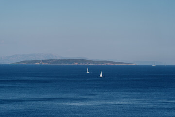 Two sailing boats glide over the calm blue sea with rolling hills in the distance, capturing the...