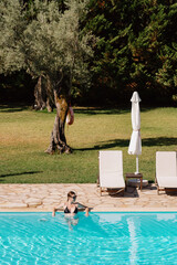 A peaceful setting featuring a woman relaxing in a clear blue swimming pool, surrounded by greenery and sun loungers.