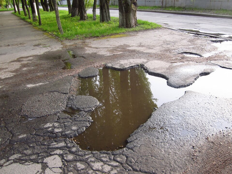 Old sidewalk pavement big cracked asphalt pothole full of spring water, broken city street asphalt road puddle, springtime new green grass and trees reflection.