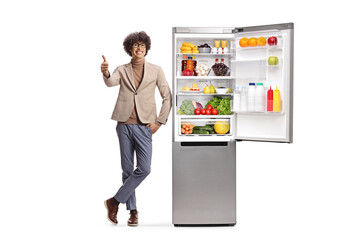 Happy young man gesturing thumbs up and standing next to a fridge