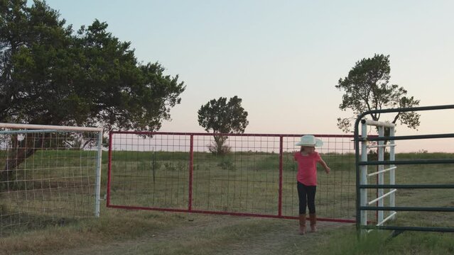 A girl with a cowboy hats closes the gate to a pasture