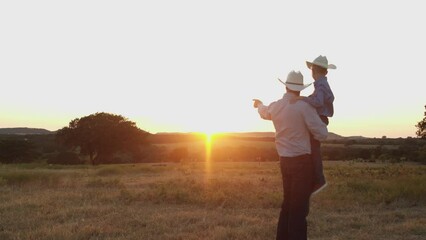 A father holds his sun as they watch the sun set in the distance on their farm