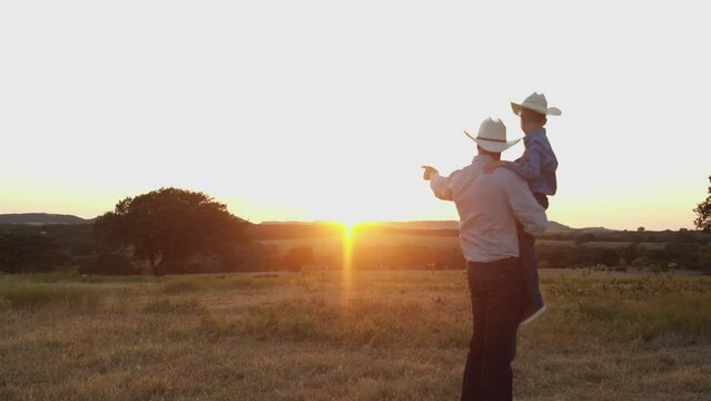 A father holds his sun as they watch the sun set in the distance on their farm