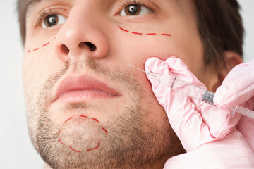 Young man with marked face receiving injection on light background, closeup. Plastic surgery concept