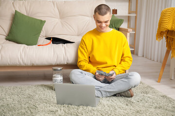 Male student sitting on floor with laptop, jar bank and money in room. Concept of savings for...