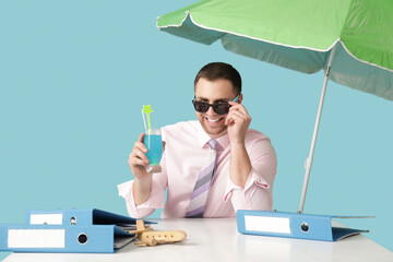 Happy male office worker in sunglasses sitting at table with cocktail on blue background. Summer...