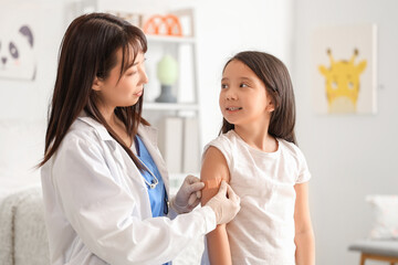 Female Asian doctor applying medical patch on little girl's arm after vaccination in clinic