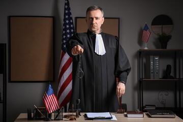 Mature judge with handcuffs and gavel at table in dark office