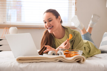 Young woman with laptop eating potato chips in bedroom - Powered by Adobe