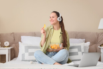 Young woman with headphones eating potato chips in bedroom