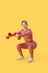 Young female fitness trainer exercising with dumbbells on yellow background