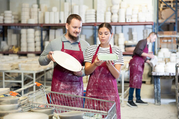 Two young pottery experts, girl and bearded man in maroon aprons, closely examining and discussing...