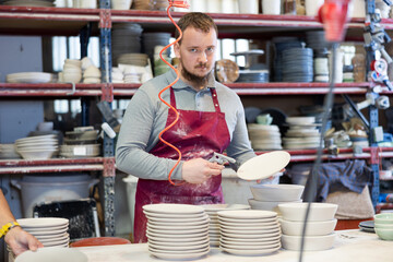 Focused bearded potter using air gun to remove dust from freshly made ceramic plates, standing at...