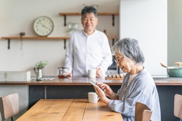 スマホを使う笑顔の高齢者女性とキッチンにいる高齢者男性
