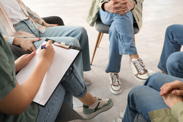 Female psychologist writing in clipboard at group therapy session, closeup