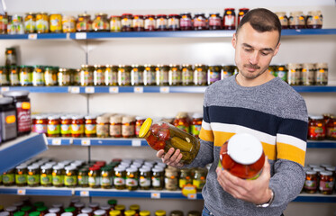 Man near display of canned vegetables in Russian goods store. Male buyer chooses jar of pickled...