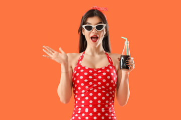 Beautiful young shocked woman in swimsuit with bottle of cold soda on orange background. Summer...