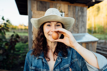 Funny Woman Farmer in denim with a fresh egg, making a funny face. A woman in hat exhibits sheer happiness with a fresh egg in hand, standing on her farm Sustainable lifestyle, organic farmer
