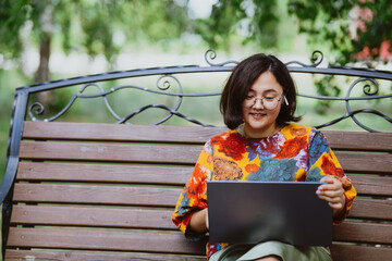 Female professional enjoys remote video conferencing on a calm sunny day in a lush green park. A cheerful adult Asian woman, engages in lively video calls on her laptop while sitting on park