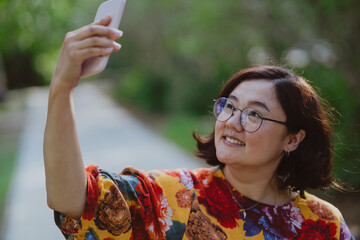 Happy woman taking a selfie with her mobile phone in a blooming park. Young entrepreneur with digital devices capturing a selfie, staying connected in an urban green space