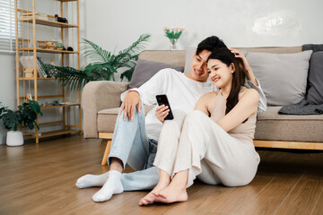 Relaxed and smiling, an Asian couple shares a close moment while looking at a smartphone, sitting on the living room floor.