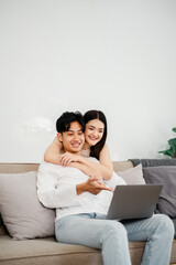 Affectionate Asian couple is cuddling on the sofa, with a laptop, creating a scene of domestic bliss and shared interests.