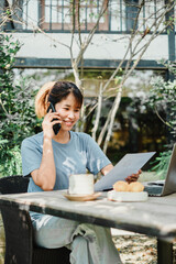 Woman talks on the phone and reviews a document at an outdoor wooden table with a laptop, surrounded by lush greenery.