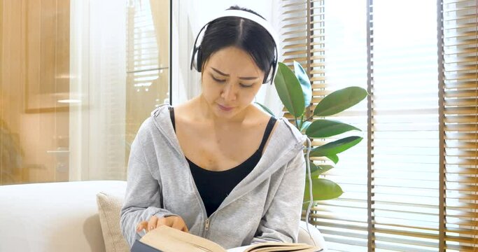 Happy asian woman use headphones listen to music reading book sitting on cozy sofa in living room at home. Female asian people work at home research book read magazine smiling happy reader lifestyle