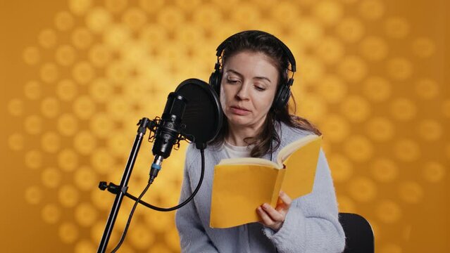 Narrator wearing headset reading aloud from book into mic against yellow background. Upbeat professional voice actor recording audiobook, creating engaging media content for listeners, camera B