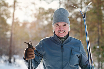 Portrait of cheerful active senior man holding skis and poles standing in forest park on cold winter day smiling at camera