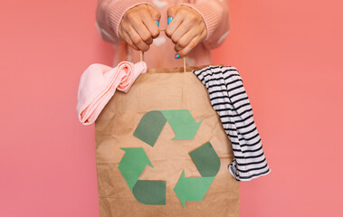 Paper bag with recycling sign with old clothes in hands against pink background.