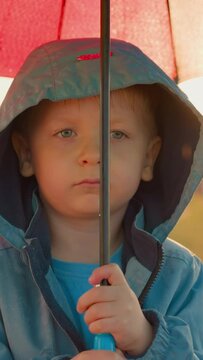 Upset kid with umbrella at rainfall on riverbank. Thoughtful little boy in bad mood stands alone in green field on rainy day. Child in discomfortable place