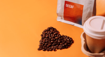 Coffee beans in the shape of a heart on an orange background next to packaging with text decaf and a paper cup in a tray.
