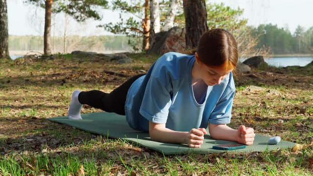 A young red-haired girl is doing outdoor sports in the forest. The girl takes a plank pose and listens to inspiring music on her phone.