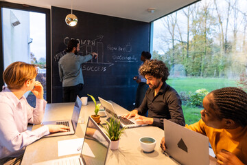 An animated business meeting unfolds with a diverse group of professionals in a well-lit, glass-walled office. One team member writes on a blackboard, illustrating growth strategies, as his colleagues