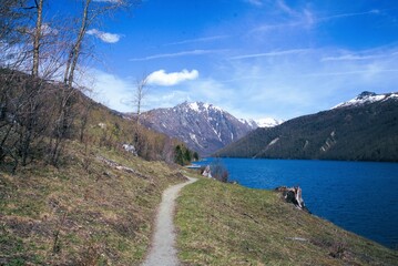 Narrow footpath winding next to large calm lake, surrounded by scrub and bare trees on the near...