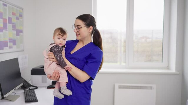 A happy brunette girl in a blue uniform a doctor in glasses holds a little baby girl in her arms and finds a common language with her during prevention in a modern clinic with a pediatrician