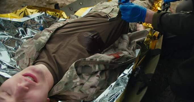 Confident male military man in a dark green uniform and with blue medical gloves cuts the T-shirt of a wounded soldier to stop bleeding during training and combat exercises providing medical care